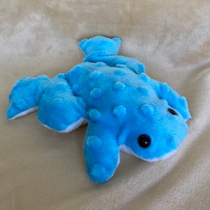 Peut inclure: Un jouet en peluche de grenouille bleu avec des accents blancs et des yeux noirs. La grenouille a une texture bosselée.
