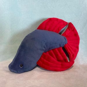 Puede incluir: Un peluche con forma de pez betta. El pez tiene un cuerpo azul y una gran cola en forma de abanico de color rojo brillante. Un pequeño botón negro está en el costado del cuerpo. El juguete es suave y parece estar hecho de tela.