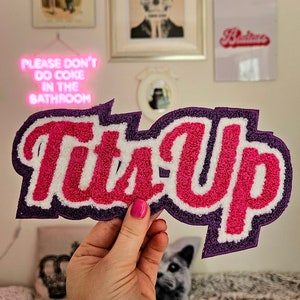 May include: A pink and white "Tits Up" patch with purple edging. The patch is made of fuzzy material and is being held by a hand.