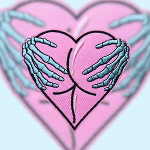 May include: A pink heart-shaped patch with two blue skeletal hands holding it. The hands are embroidered in a light blue colour.