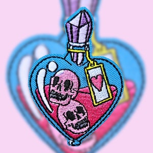May include: A heart-shaped glass bottle with a purple crystal stopper, filled with a pink liquid and two pink skulls. The bottle has a tag with a red heart on it.