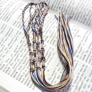 May include: Braided friendship bracelets in various colors, including blue, brown, and beige, are displayed open on a book. The bracelets are made of thin, woven threads and are knotted together. The book's pages are visible.