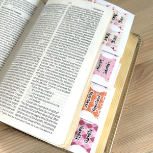 May include: An open book with gold-edged pages featuring printed text and a series of decorative adhesive tabs attached to the side. The tabs display the words Grace, Salvation, Repentance, and Humility in various patterns including pink and orange.