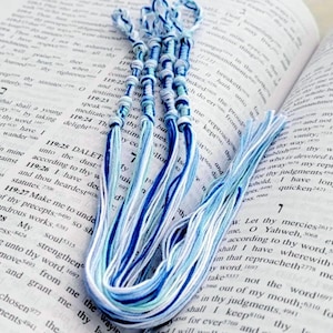 May include: A handmade bookmark crafted from multiple strands of embroidery floss in shades of navy blue, sky blue, and white. The design features a series of intricate knots at the top, transitioning into long, loose, flowing fringe at the bottom.
