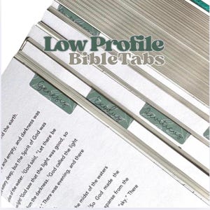 May include: A set of sage green low profile Bible tabs attached to the edges of book pages. The tabs feature white cursive text labeling the books of the Bible, including Genesis, Exodus, and Leviticus. The text Low Profile Bible Tabs is overlaid.