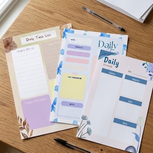 Può includere: Tre blocchi per la pianificazione giornaliera colorati con accenti floreali e testo. Uno recita "Daily Task List" con sezioni per "Urgent (Do it)" e "Today's Motivation". Un altro dice "Daily Planner" con sezioni per "Schedule" e "Goals". Una penna è appoggiata sulla superficie di legno.