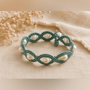 May include: A bracelet crafted from teal-colored cord featuring a repeating macrame knot pattern that forms oval shapes. Each oval contains a single round, white pearl. The bracelet includes a small knotted closure at the end of the band.