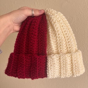 May include: A handmade crochet beanie in a two-tone design. One half is a deep red, the other is cream-colored. The hat is made with a ribbed stitch pattern and has a folded brim. This winter hat is perfect for cold weather.