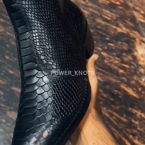 Puede incluir: Bota de cuero negro con un patrón texturizado similar a la piel de serpiente. La bota tiene una punta afilada y un tacón bajo. El texto "POWER_KNOTS" es visible en el lateral. La bota es sostenida por una persona.