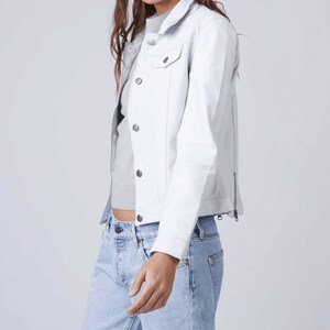 May include: A white denim jacket with a zipper on the side and a button closure. The jacket has a pocket on the left chest.