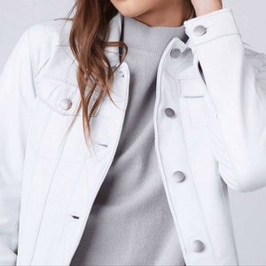May include: A white leather jacket with silver buttons. The jacket has a classic denim style with a pointed collar and flap pockets.