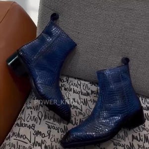 Peut inclure: Une paire de bottines bleues avec un motif texturé en forme de crocodile. Les bottines ont un bout pointu et un petit talon noir. Les bottines reposent sur une surface à motifs.