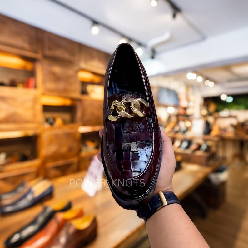 May include: A burgundy leather loafer with a gold chain detail. The shoe has a textured, crocodile-like pattern and a rounded toe. The sole is black. The text "POWER KNOTS" is visible on the shoe.