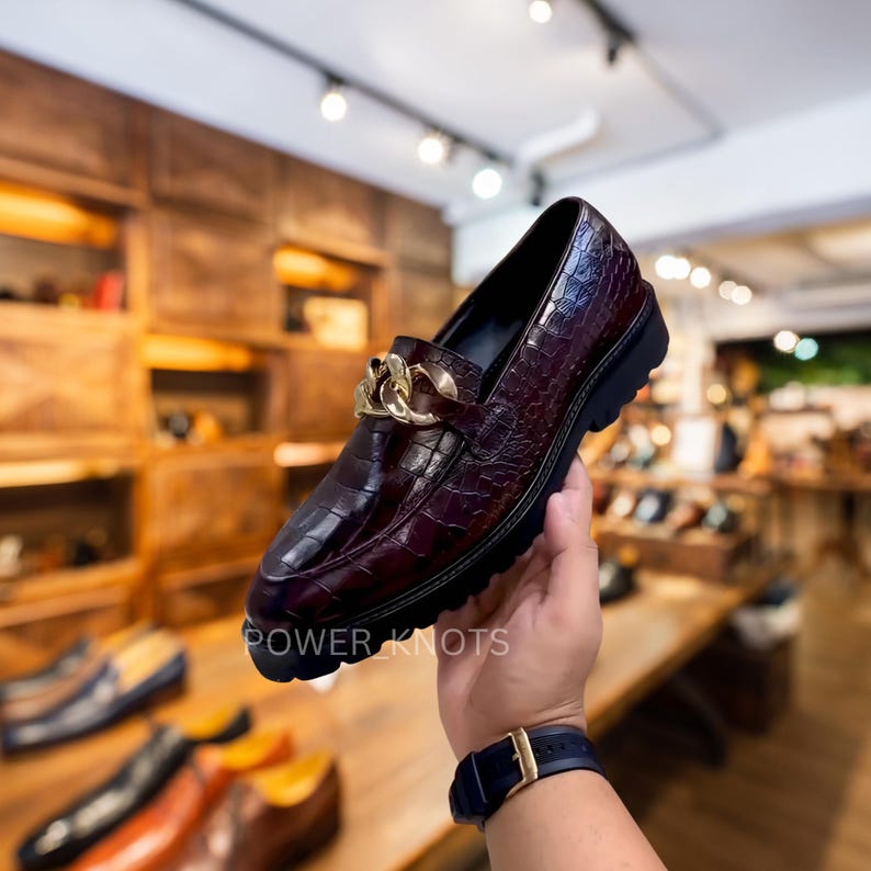 May include: A burgundy leather loafer with a crocodile texture and a gold chain detail. The shoe has a thick black sole and the text "POWER KNOTS" is visible.