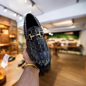 May include: Black leather loafer with a crocodile texture, gold-coloured buckle, and studded trim. The shoe has a classic design with a modern edge. The text "POWER_KNOTS" is visible on the side of the shoe.