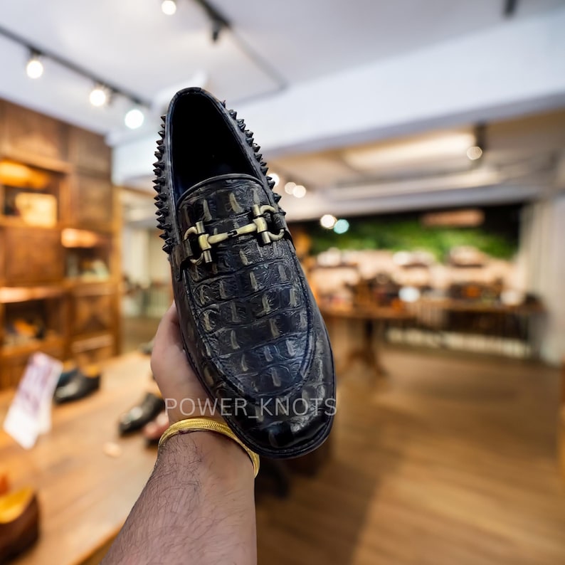 May include: Black leather loafer with a crocodile texture, gold-coloured buckle, and studded trim. The shoe has a classic design with a modern edge. The text "POWER_KNOTS" is visible on the side of the shoe.