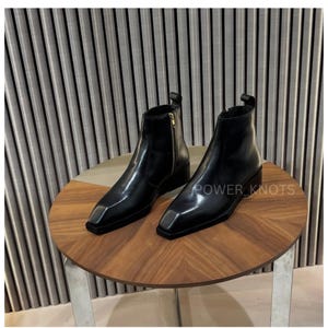 May include: A pair of black leather ankle boots with square toes and a side zipper. The boots are displayed on a round wooden table with a silver base. The text "POWER_KNOTS" is visible on the table.