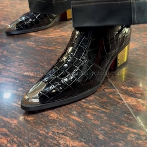 May include: A black ankle boot with a glossy crocodile-embossed texture, featuring a pointed toe with a polished silver-tone metal cap, a block heel with a gold-tone metal accent, and a red sole, worn with dark trousers.