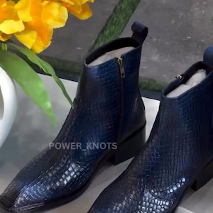 Puede incluir: Botines azul marino con textura de efecto cocodrilo. Tienen punta cuadrada, cremallera lateral y tacón pequeño. El texto "POWER_KNOTS" es visible. Flores amarillas en el fondo. Calzado elegante para diversas ocasiones.
