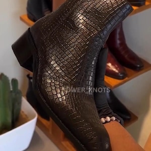 Puede incluir: Botín marrón oscuro con un patrón texturizado similar al de un reptil. El botín tiene un tacón cuadrado y una puntera en punta. Otras botas son visibles en una estantería de madera en el fondo. El texto "POWER_KNOTS" es visible.