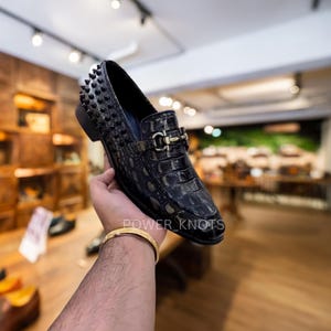 May include: Black leather loafer with a crocodile texture, featuring black spikes on the heel and a gold-coloured buckle. The shoe is held up in front of a store background. The text "POWER KNOTS" is visible on the shoe.