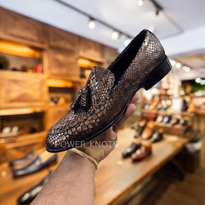 May include: A brown and gold loafer with a snakeskin pattern. The shoe has a tassel detail and a black sole. The text "POWER_KNOTS" is visible on the shoe. The shoe is held up in front of a wooden background.