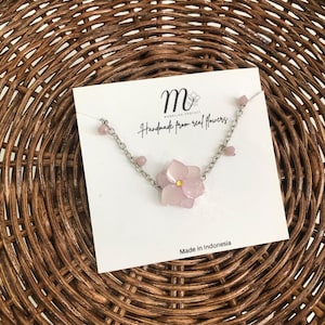 May include: A necklace with a light pink flower pendant and a silver chain. The card reads "Handmade from real flowers" and "Made in Indonesia". The necklace is displayed on a woven wicker surface. The pendant has a yellow center.