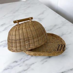 May include: A woven rattan serving dish with a domed lid and a wooden handle. The dish is light brown and sits on a white marble surface. The lid is slightly ajar, revealing the interior of the dish.