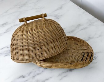 Puede incluir: Un plato para servir de ratán tejido de color marrón claro, con una tapa abovedada y un asa de madera. El plato está sobre una superficie de mármol blanco. La tapa está ligeramente abierta, mostrando el interior.