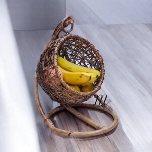 Puede incluir: Una cesta de frutas de ratán tejido marrón con diseño de silla colgante. La cesta está llena de plátanos amarillos y una fruta de color naranja rosado. La cesta tiene una base circular y un asa en la parte superior.