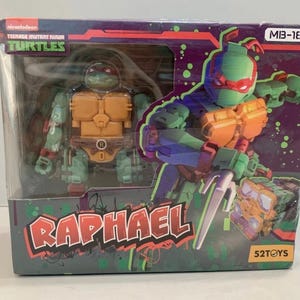 Può includere: Un action figure di Raphael delle Tartarughe Ninja in una scatola di cartone rettangolare. La confezione presenta un'illustrazione del personaggio verde e arancione, il nome RAPHAEL in lettere rosse e il logo 52TOYS su sfondo viola.