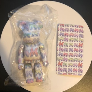 May include: A colorful, cartoon-style bear figurine with a repeating pattern of faces. The bear is packaged in a clear plastic bag and comes with a card featuring the same pattern. The card has the text "© KAI TANAKA" printed on the bottom.