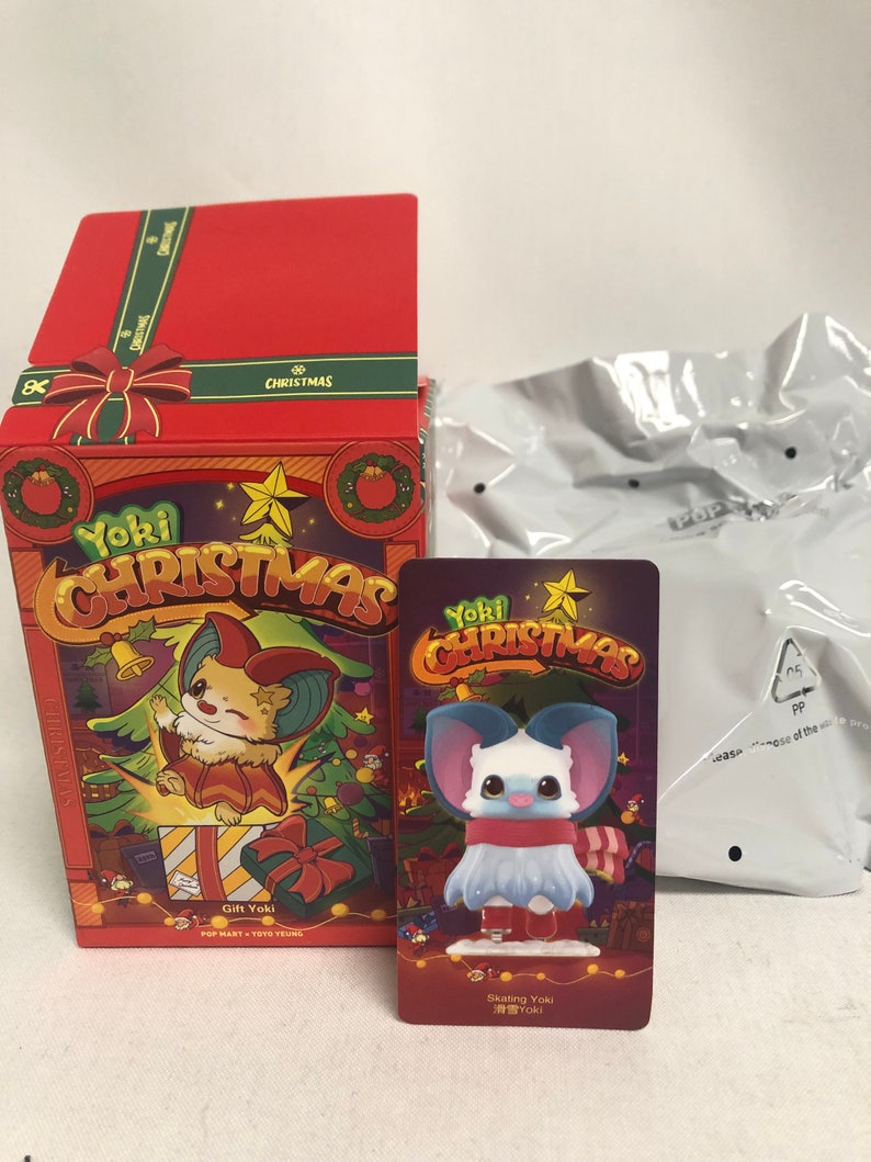 Puede incluir: Una caja de regalo roja y una tarjeta con un personaje de dibujos animados con orejas grandes, parte de la colecci&oacute;n "Yoki Christmas". La tarjeta muestra una figura con una bufanda roja y patines. Tambi&eacute;n hay una bolsa de pl&aacute;stico en la imagen.