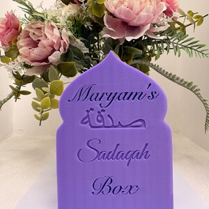 Puede incluir: Una caja Sadaqah de color lila con el nombre "Maryam's", escritura &aacute;rabe y las palabras "Sadaqah Box". La caja est&aacute; frente a un ramo de flores rosas y blancas.