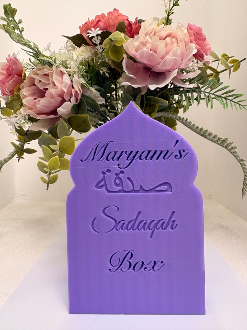 Puede incluir: Una caja Sadaqah de color lila con el nombre "Maryam's", escritura &aacute;rabe y las palabras "Sadaqah Box". La caja est&aacute; frente a un ramo de flores rosas y blancas.