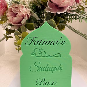 Puede incluir: Una caja Sadaqah verde con forma de arco, con el nombre "Fatima's" y la palabra "Sadaqah" en &aacute;rabe e ingl&eacute;s. La caja est&aacute; decorada con un ramo de flores rosas y blancas.