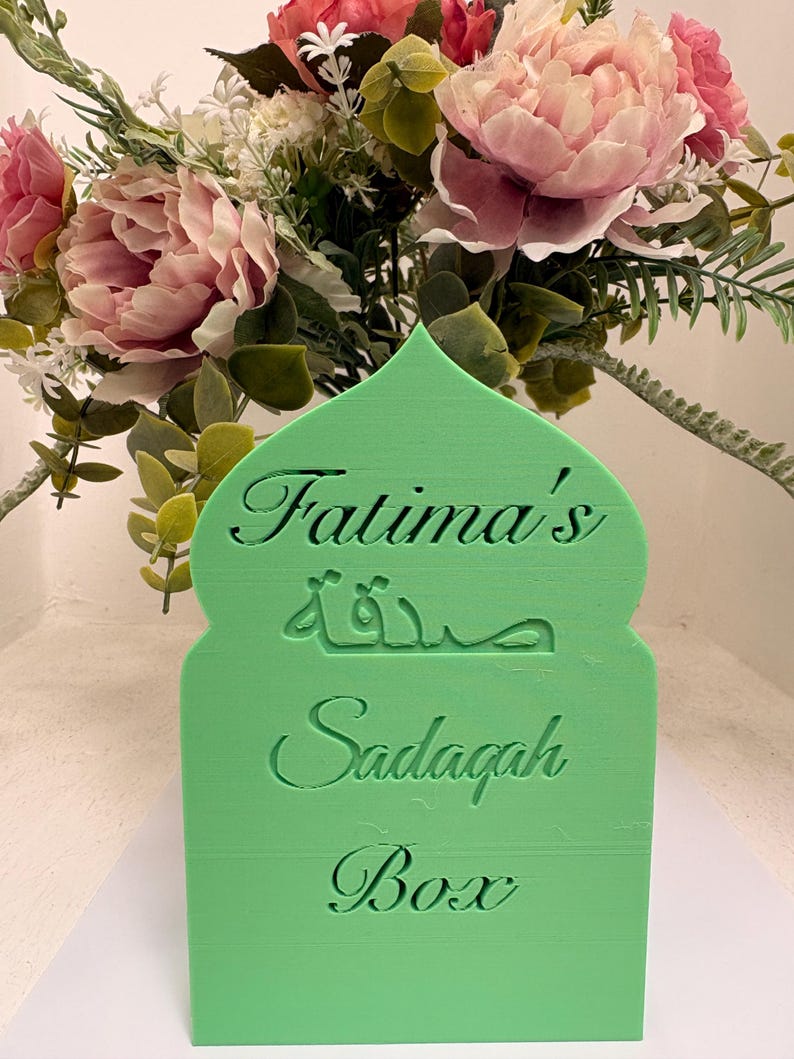 Puede incluir: Una caja Sadaqah verde con forma de arco, con el nombre "Fatima's" y la palabra "Sadaqah" en &aacute;rabe e ingl&eacute;s. La caja est&aacute; decorada con un ramo de flores rosas y blancas.