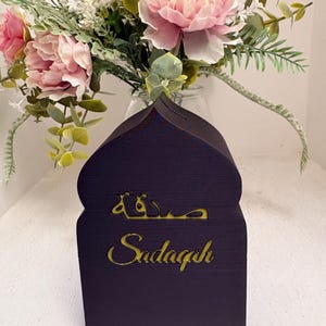 Puede incluir: Una caja Sadaqah de madera azul oscuro con una parte superior en forma de mezquita. La palabra "Sadaqah" est&aacute; escrita en letras doradas. Un ramo de flores rosas y blancas se encuentra en un jarr&oacute;n de cristal en el fondo.
