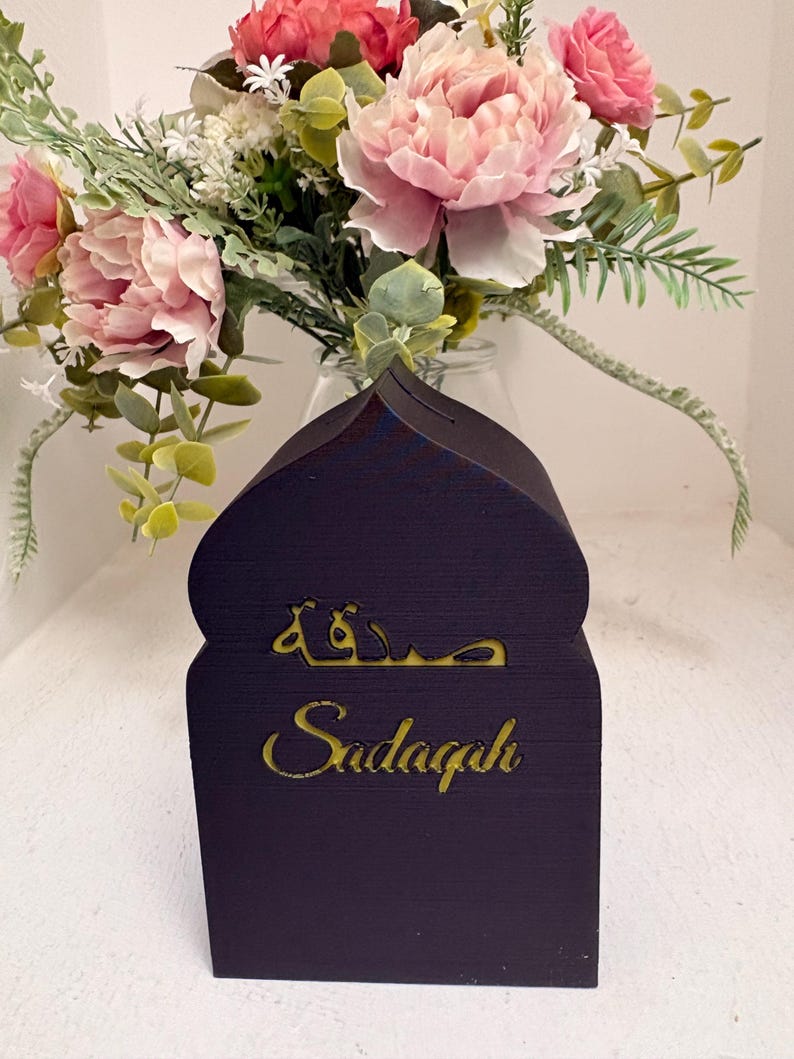 Puede incluir: Una caja Sadaqah de madera azul oscuro con una parte superior en forma de mezquita. La palabra "Sadaqah" est&aacute; escrita en letras doradas. Un ramo de flores rosas y blancas se encuentra en un jarr&oacute;n de cristal en el fondo.