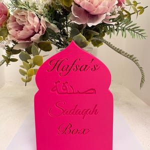 Puede incluir: Una caja Sadagah de color rosa brillante con el nombre "Hafsa's" y las palabras "Sadagah Box" en ingl&eacute;s y &aacute;rabe. La caja est&aacute; frente a un jarr&oacute;n con flores rosas y blancas.