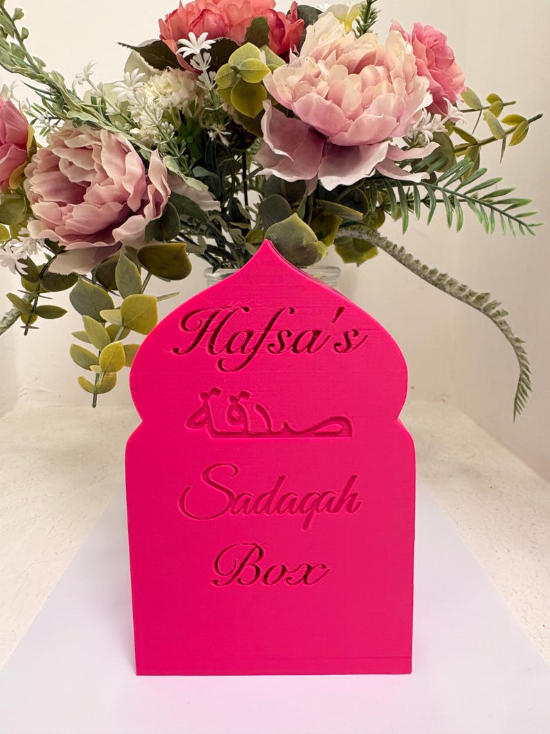 Puede incluir: Una caja Sadagah de color rosa brillante con el nombre "Hafsa's" y las palabras "Sadagah Box" en ingl&eacute;s y &aacute;rabe. La caja est&aacute; frente a un jarr&oacute;n con flores rosas y blancas.