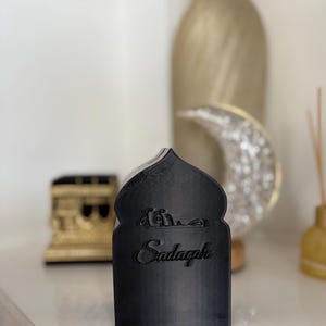 Puede incluir: Una pieza decorativa negra con una parte superior arqueada y la palabra "Sadaqah" en escritura cursiva. La pieza tiene un dise&ntilde;o isl&aacute;mico, con escritura &aacute;rabe encima de la palabra. Otros elementos decorativos est&aacute;n en el fondo.