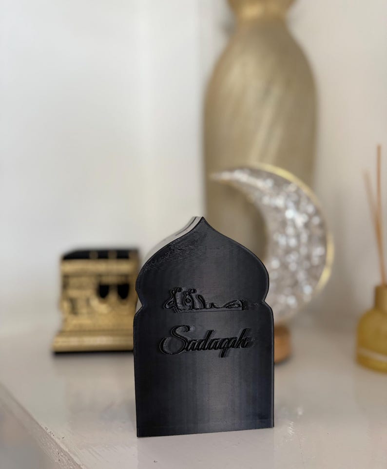 Puede incluir: Una pieza decorativa negra con una parte superior arqueada y la palabra "Sadaqah" en escritura cursiva. La pieza tiene un dise&ntilde;o isl&aacute;mico, con escritura &aacute;rabe encima de la palabra. Otros elementos decorativos est&aacute;n en el fondo.