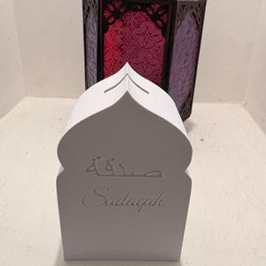 Puede incluir: Una caja Sadagah blanca con forma de mezquita con la palabra "Sadagah" y escritura &aacute;rabe. Una linterna decorativa de metal oscuro con vidrieras rojas y moradas en el fondo.