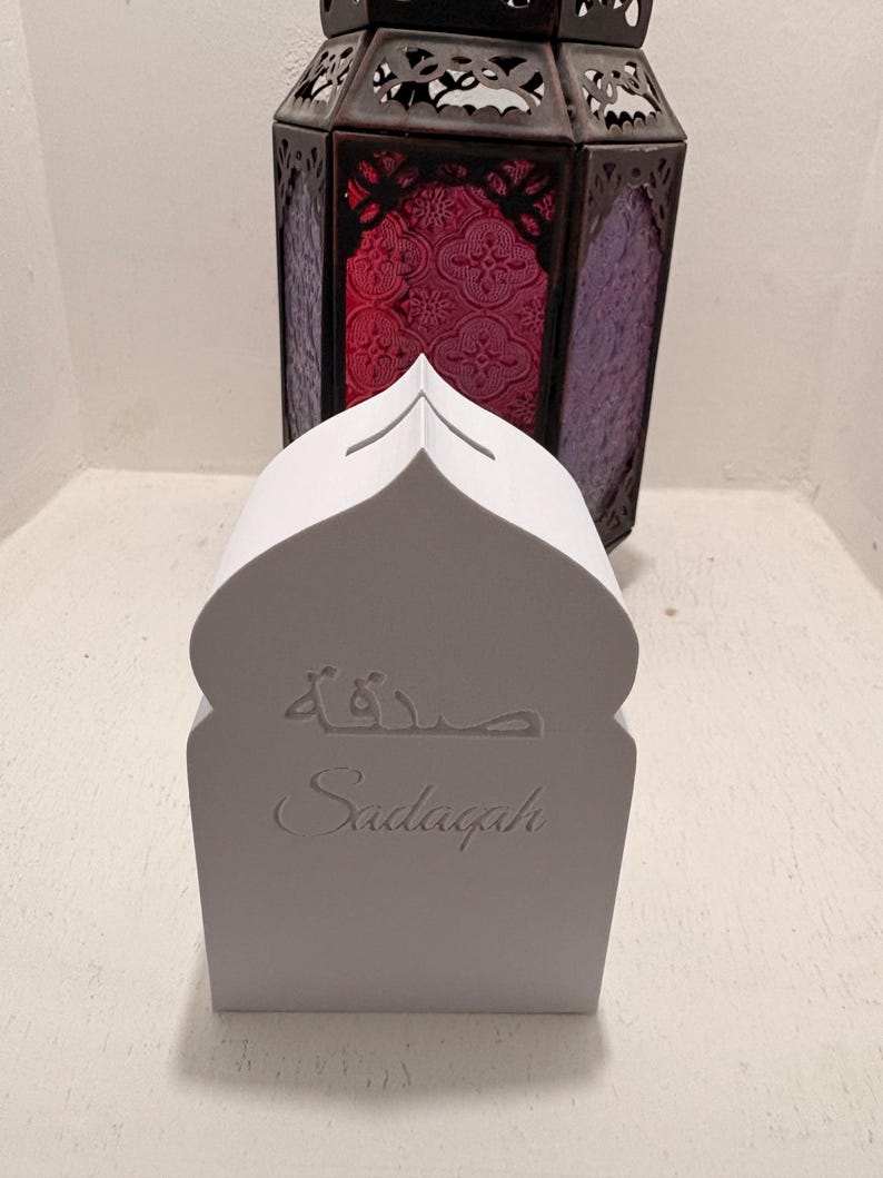 Puede incluir: Una caja Sadagah blanca con forma de mezquita con la palabra "Sadagah" y escritura &aacute;rabe. Una linterna decorativa de metal oscuro con vidrieras rojas y moradas en el fondo.