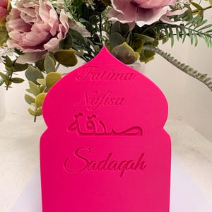 Puede incluir: Un letrero decorativo rosa brillante, cortado con l&aacute;ser, con una parte superior arqueada. El letrero presenta los nombres "Fatima" y "Nafisa" en cursiva, escritura &aacute;rabe y la palabra "Sadaqah".