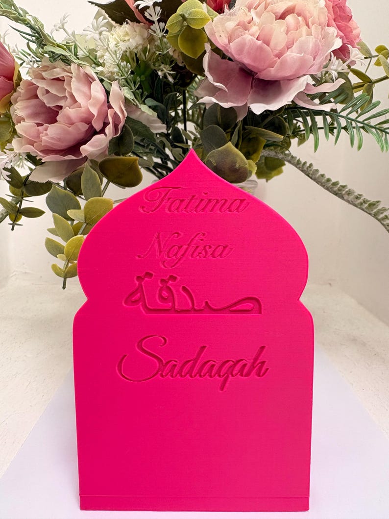 Puede incluir: Un letrero decorativo rosa brillante, cortado con l&aacute;ser, con una parte superior arqueada. El letrero presenta los nombres "Fatima" y "Nafisa" en cursiva, escritura &aacute;rabe y la palabra "Sadaqah".