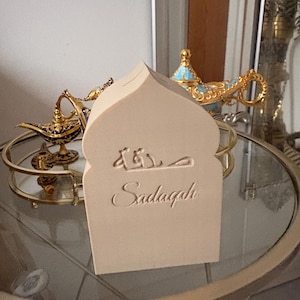 Puede incluir: Una caja Sadaqah beige, con forma de arco, con escritura &aacute;rabe y la palabra inglesa "Sadaqah" grabada en la parte delantera. La caja est&aacute; sobre una mesa con tapa de cristal y ribetes dorados, con otros objetos decorativos visibles al fondo.