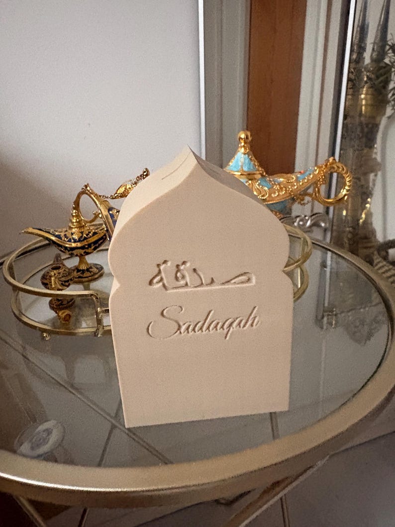 Puede incluir: Una caja Sadaqah beige, con forma de arco, con escritura &aacute;rabe y la palabra inglesa "Sadaqah" grabada en la parte delantera. La caja est&aacute; sobre una mesa con tapa de cristal y ribetes dorados, con otros objetos decorativos visibles al fondo.