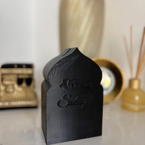 Puede incluir: Caja decorativa negra con una parte superior arqueada y la palabra "Sadaqah" grabada en la parte delantera. La caja tiene un dise&ntilde;o similar a una mezquita y probablemente se utiliza para donaciones ben&eacute;ficas. Otros elementos decorativos est&aacute;n en el fondo.